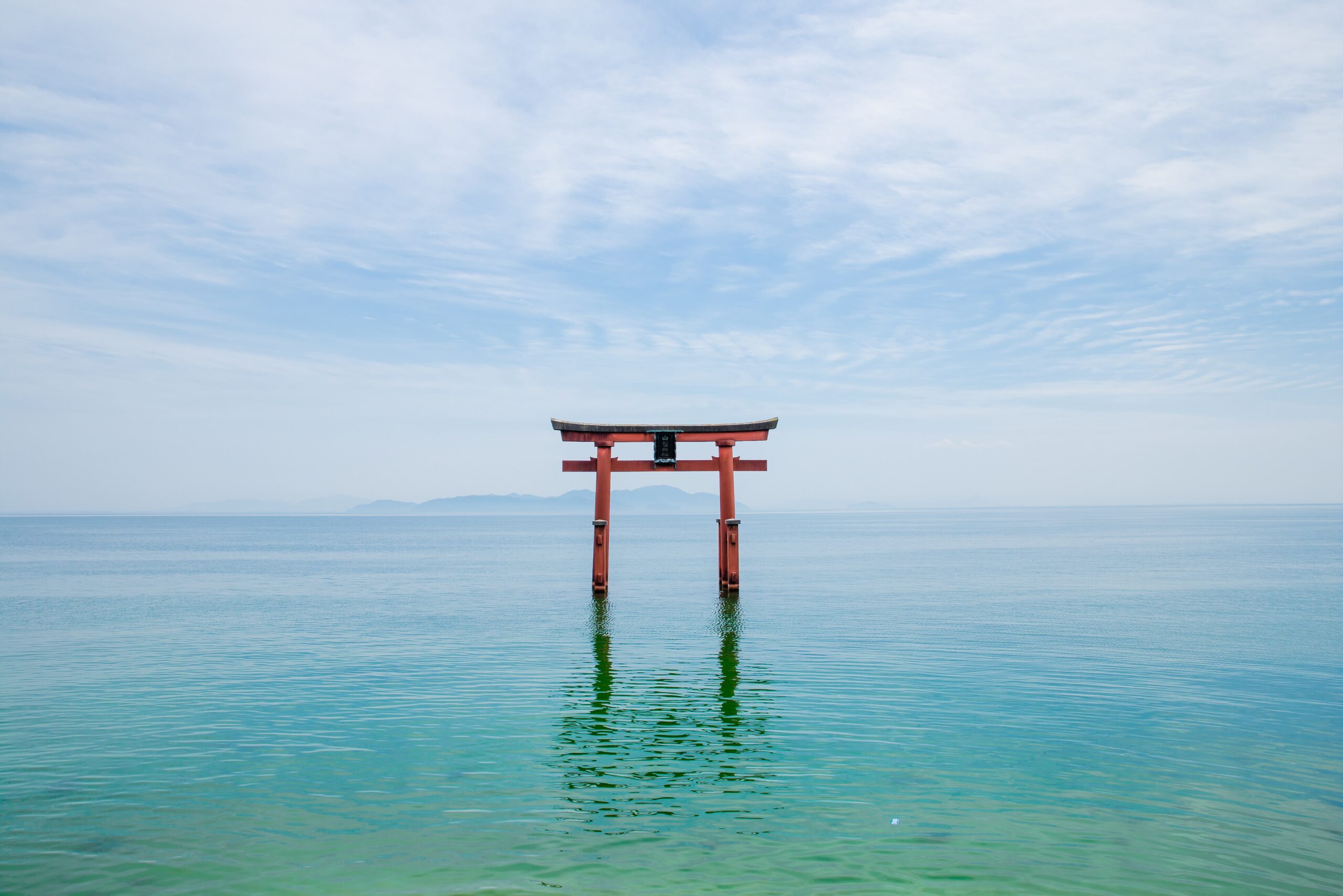 Red torii gate standing in calm turquoise sea under a blue, cloudy sky.