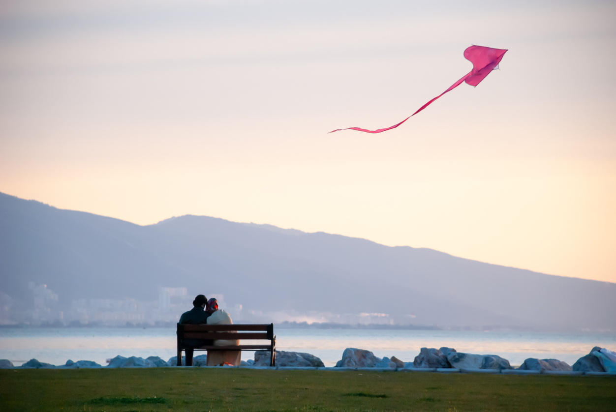 A couple sitting close together on a park bench by the water at sunset, with a pink kite flying overhead and soft hills in the distance.